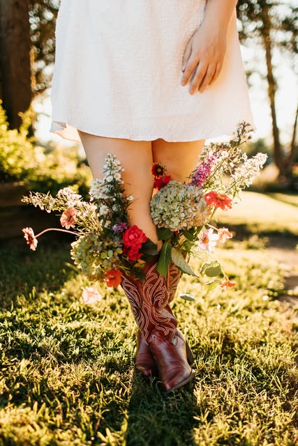 Cowboy boots filled with colorful flowers beside a white dress in golden light