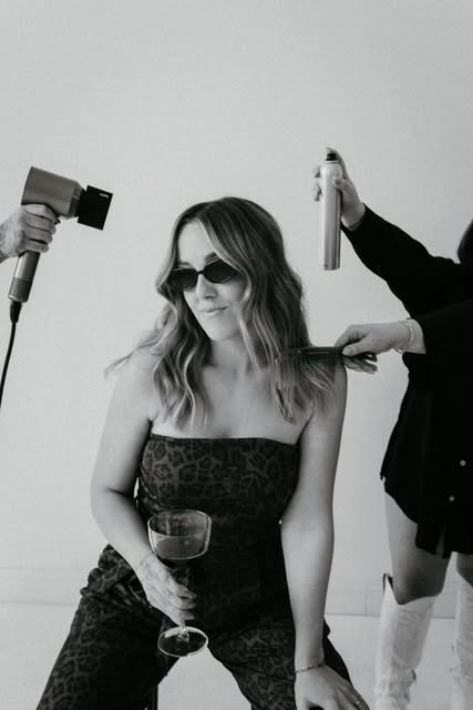 Black-and-white portrait of a woman in sunglasses enjoying a drink while her hair is styled