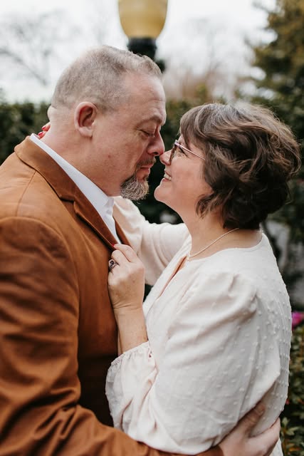 A couple embracing closely outdoors, wearing a brown jacket and a white blouse